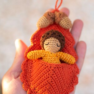 May include: A crocheted doll with brown hair and closed eyes is sleeping in a crocheted orange leaf bed. The bed is decorated with two small crocheted acorns.