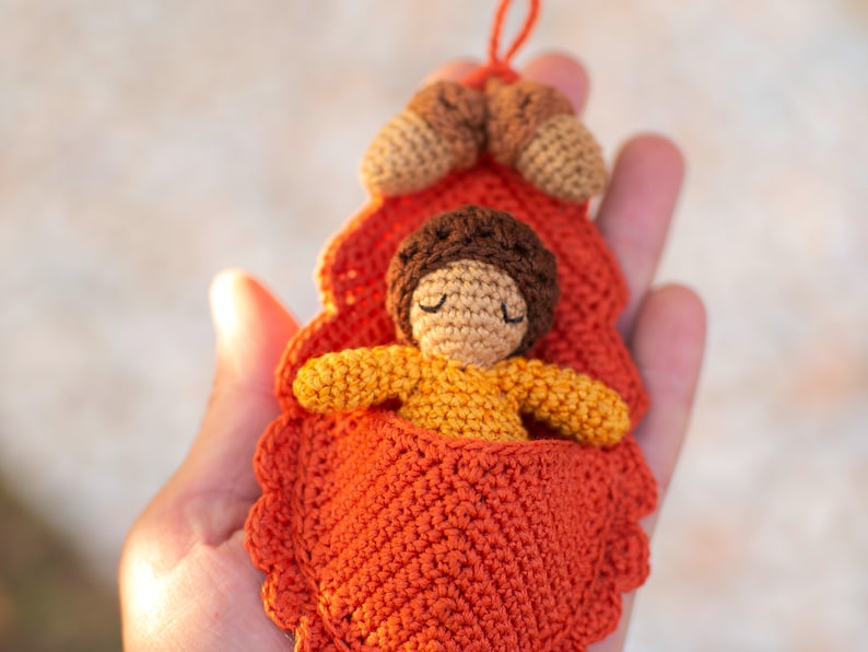 May include: A crocheted doll with brown hair and closed eyes is sleeping in a crocheted orange leaf bed. The bed is decorated with two small crocheted acorns.