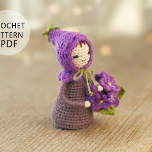 Könnte beinhalten: Häkelmuster-PDF für eine lila gehäkelte Puppe, die einen Strauß lila Blumen hält. Die Puppe trägt einen lila Kapuzenhut und hat einen süßen Gesichtsausdruck.