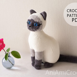 May include: A crocheted Siamese cat toy with cream-colored fur, dark gray ears, face, paws, and tail, and bright blue eyes. Next to it is a small blue glass vase with a single pink rose. The text CROCHET PATTERN PDF and AniAmiCrochet are visible.