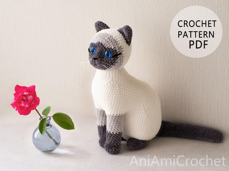 May include: A crocheted Siamese cat toy with cream-colored fur, dark gray ears, face, paws, and tail, and bright blue eyes. Next to it is a small blue glass vase with a single pink rose. The text CROCHET PATTERN PDF and AniAmiCrochet are visible.