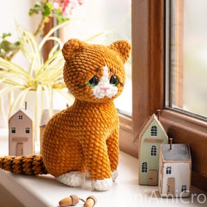 May include: A crocheted orange and white cat plush toy with large dark eyes, a pink nose, and a striped tail. The cat is sitting upright, featuring a textured yarn finish, white paws, and a white patch on its face. The text AniAmiCrochet is visible.