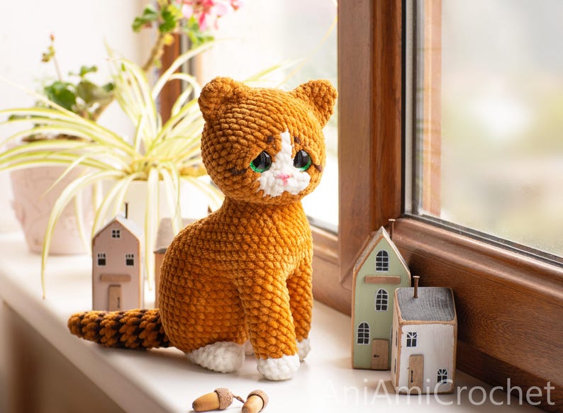 May include: A crocheted orange and white cat plush toy with large dark eyes, a pink nose, and a striped tail. The cat is sitting upright, featuring a textured yarn finish, white paws, and a white patch on its face. The text AniAmiCrochet is visible.
