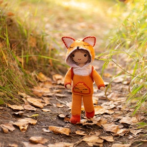 May include: A crocheted fox doll wearing an orange and white hooded jumpsuit. The doll is standing on a path covered in fallen leaves.