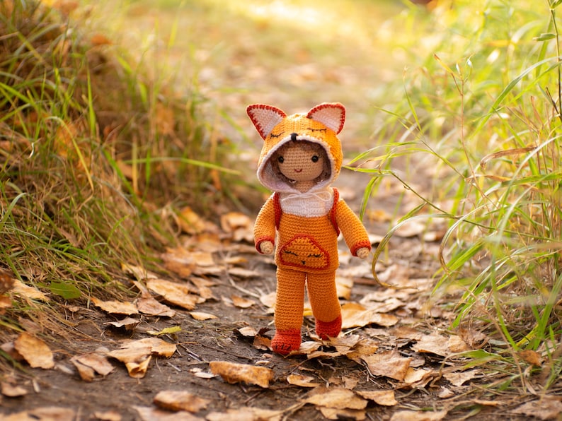 May include: A crocheted fox doll wearing an orange and white hooded jumpsuit. The doll is standing on a path covered in fallen leaves.