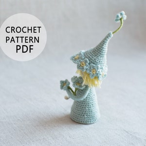 Könnte beinhalten: Eine gehäkelte Puppe mit einem blauen Hut mit gelben Blumen und einem Strauß blauer Blumen in der Hand. Die Puppe steht auf einer weißen Oberfläche. CROCHET PATTERN PDF.