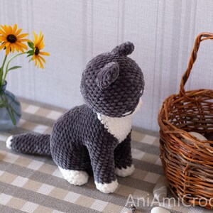 May include: A dark gray and white crocheted cat plush toy with a textured yarn finish, featuring white paws and a white chest patch. The text AniAmiCrochet is visible in the bottom right corner of the image, indicating the creator of the item.