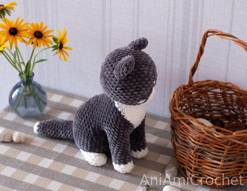 May include: A dark gray and white crocheted cat plush toy with a textured yarn finish, featuring white paws and a white chest patch. The text AniAmiCrochet is visible in the bottom right corner of the image, indicating the creator of the item.