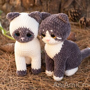 May include: Two crocheted cat plushies made from soft, textured yarn. The left cat is cream-colored with dark brown ears, face, and paws. The right cat is dark gray with a white chest, face, and paws. Both have large, glossy eyes and stitched noses.