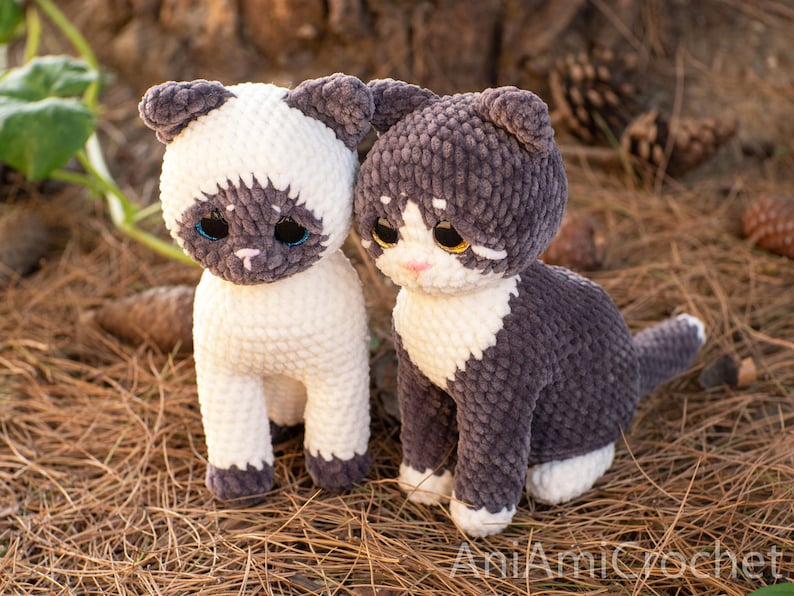 May include: Two crocheted cat plushies made from soft, textured yarn. The left cat is cream-colored with dark brown ears, face, and paws. The right cat is dark gray with a white chest, face, and paws. Both have large, glossy eyes and stitched noses.