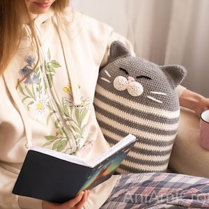 May include: A woman is sitting on a couch with a gray and white striped crocheted cat pillow. She is holding a book and a pink mug.