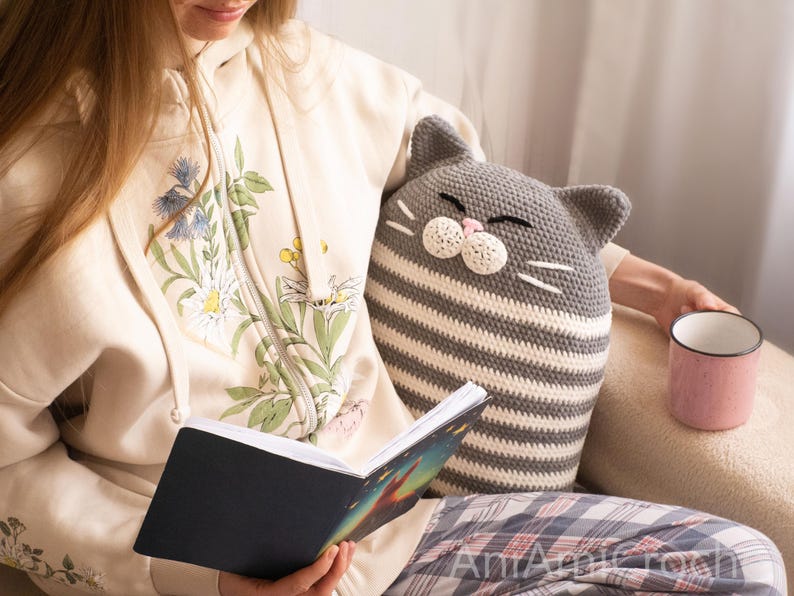 May include: A woman is sitting on a couch with a gray and white striped crocheted cat pillow. She is holding a book and a pink mug.