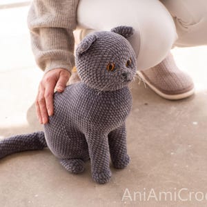 May include: A handmade, gray crocheted cat with orange eyes and a long tail. The plush cat is sitting upright, with a person's hand gently resting on its back. The cat is made of soft, textured yarn.