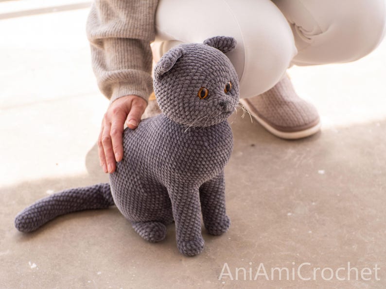 May include: A handmade, gray crocheted cat with orange eyes and a long tail. The plush cat is sitting upright, with a person's hand gently resting on its back. The cat is made of soft, textured yarn.