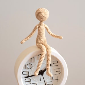 K&ouml;nnte beinhalten: Eine geh&auml;kelte Puppenfigur sitzt auf einem wei&szlig;en Wecker mit schwarzen Zahlen. Die Puppe ist beige und hat ein schlichtes Design.
