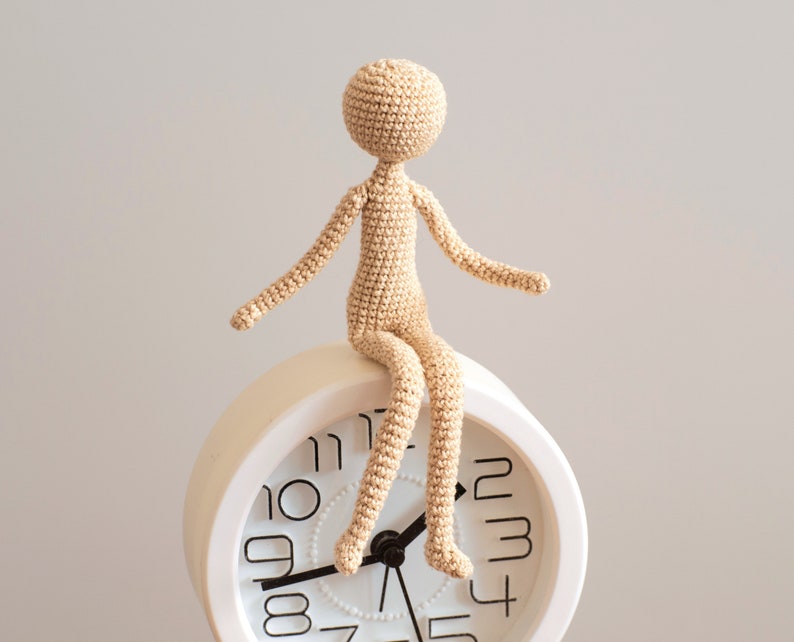 K&ouml;nnte beinhalten: Eine geh&auml;kelte Puppenfigur sitzt auf einem wei&szlig;en Wecker mit schwarzen Zahlen. Die Puppe ist beige und hat ein schlichtes Design.