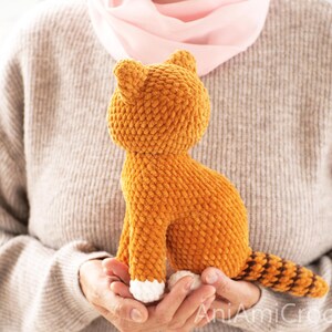 May include: A small, orange, crocheted cat plush toy with a textured, yarn-based surface. The cat features white paws, pointed ears, and a long, striped tail with alternating orange and dark brown bands. The text AniAmiCrochet is visible.