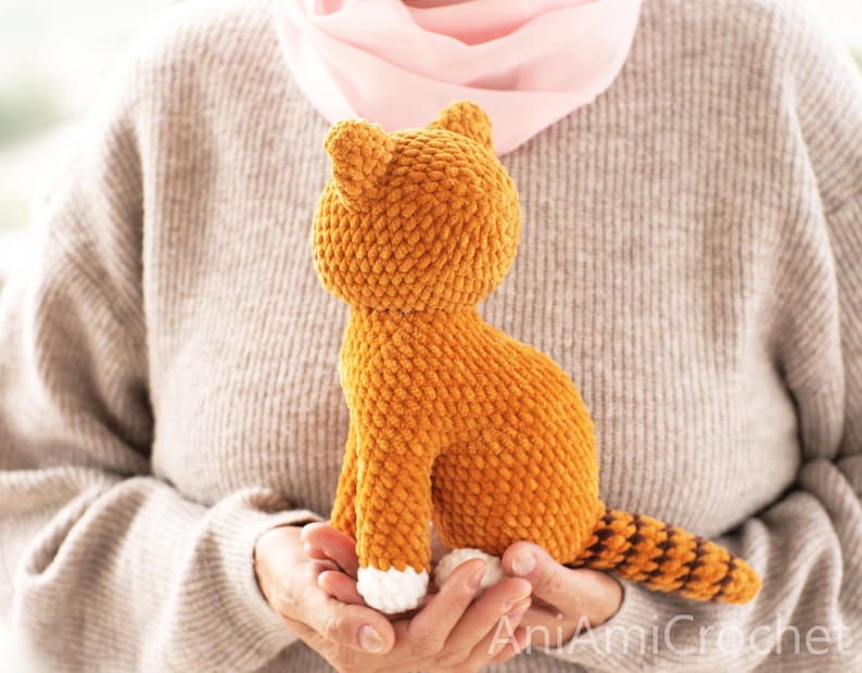 May include: A small, orange, crocheted cat plush toy with a textured, yarn-based surface. The cat features white paws, pointed ears, and a long, striped tail with alternating orange and dark brown bands. The text AniAmiCrochet is visible.