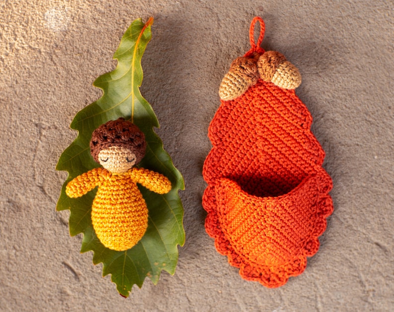 May include: A crocheted acorn doll with brown hair and a yellow dress is lying on a green leaf. A crocheted orange acorn shaped pouch with a brown top is hanging on a brown background.
