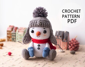Puede incluir: Patrón de crochet PDF para un adorable juguete amigurumi de muñeco de nieve. El muñeco de nieve lleva un gorro gris con un pompón, una bufanda roja y guantes azules.