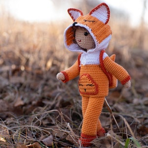 May include: A crocheted fox doll wearing a hooded orange and white jumpsuit with a backpack. The doll has a smiling face and is standing on a bed of brown leaves.