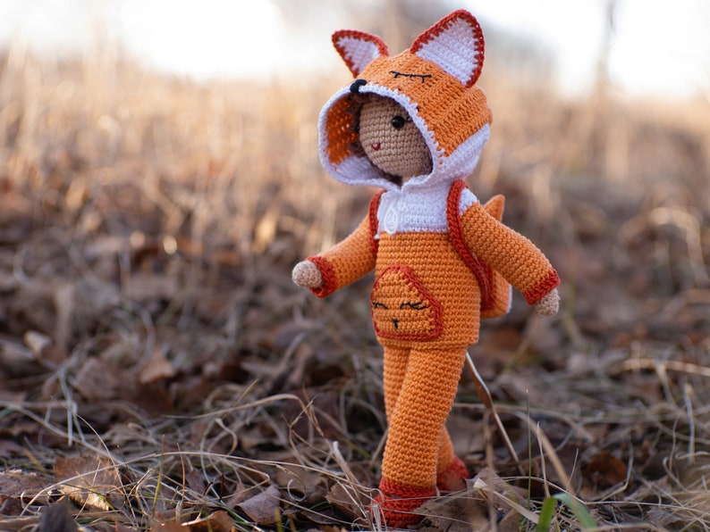 May include: A crocheted fox doll wearing a hooded orange and white jumpsuit with a backpack. The doll has a smiling face and is standing on a bed of brown leaves.