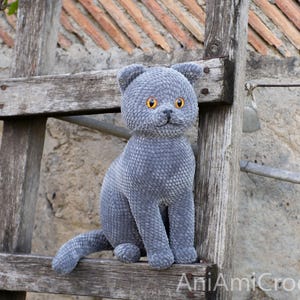 May include: A handmade, gray plush cat with bright orange eyes sits on a rustic wooden ladder. The cat has a textured, velvety appearance and is posed in a seated position. The cat's ears are perked up, and its tail is extended.