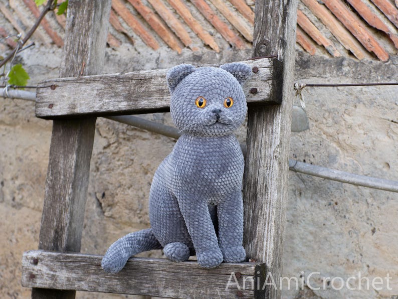 May include: A handmade, gray plush cat with bright orange eyes sits on a rustic wooden ladder. The cat has a textured, velvety appearance and is posed in a seated position. The cat's ears are perked up, and its tail is extended.