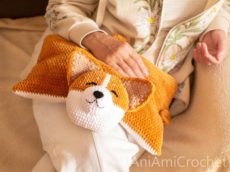 Peut inclure: Un coussin au crochet en peluche, de couleur brun dor&eacute; et blanc, en forme de chien Corgi. Le coussin pr&eacute;sente une t&ecirc;te de Corgi rembourr&eacute;e et d&eacute;taill&eacute;e avec des yeux brod&eacute;s, un nez noir et des oreilles textur&eacute;es sur un corps rectangulaire.