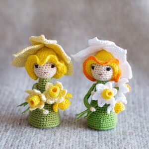 May include: Two crocheted dolls dressed as daffodils. The dolls are wearing yellow and white crocheted hats and holding bouquets of yellow and white crocheted flowers.