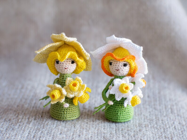 May include: Two crocheted dolls dressed as daffodils. The dolls are wearing yellow and white crocheted hats and holding bouquets of yellow and white crocheted flowers.