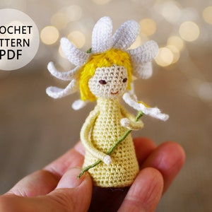 May include: Crocheted daisy doll with yellow hair and a white dress holding a green stem with a white daisy. The doll is being held in a hand. The image includes the text "CROCHET PATTERN PDF".