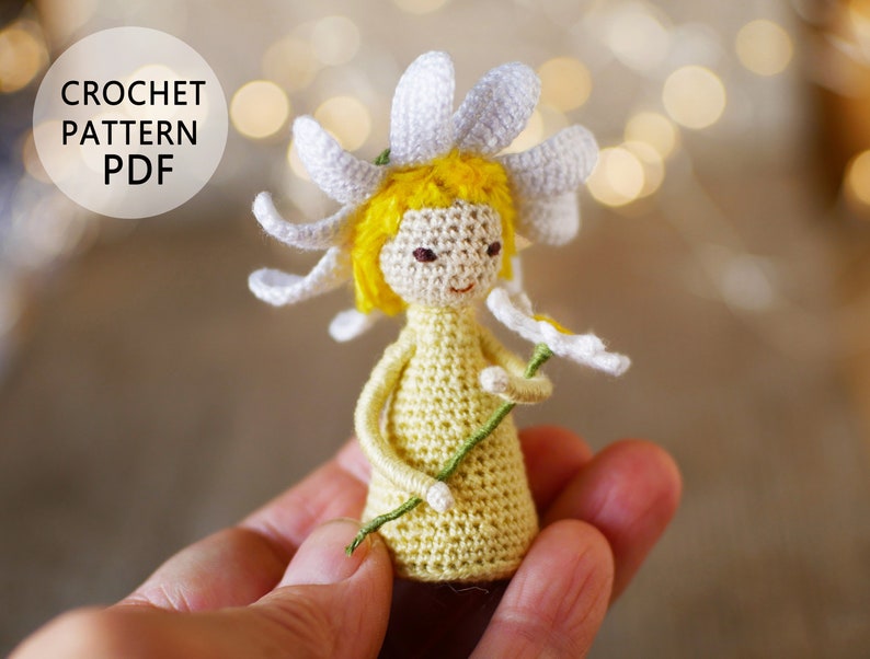 May include: Crocheted daisy doll with yellow hair and a white dress holding a green stem with a white daisy. The doll is being held in a hand. The image includes the text "CROCHET PATTERN PDF".