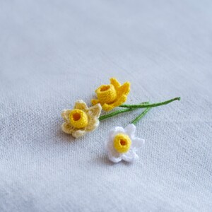 May include: Three crocheted daffodils, one white and two yellow, with green stems.