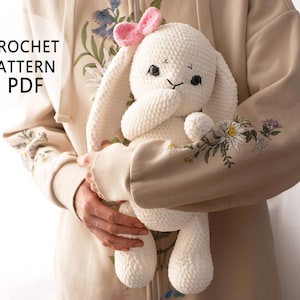May include: A white crocheted bunny with pink ears and a pink bow. The bunny is being held by a person wearing a beige hoodie with floral embroidery.