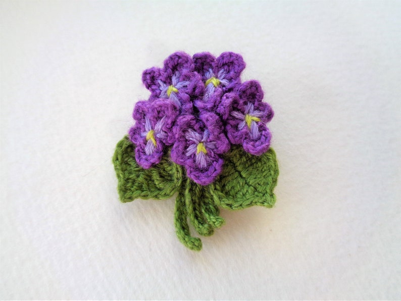 May include: A crocheted purple flower bouquet with green leaves. The flowers have yellow centers.
