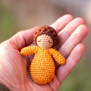 May include: A small crocheted doll with brown hair and a yellow dress. The doll is sitting in the palm of a hand.