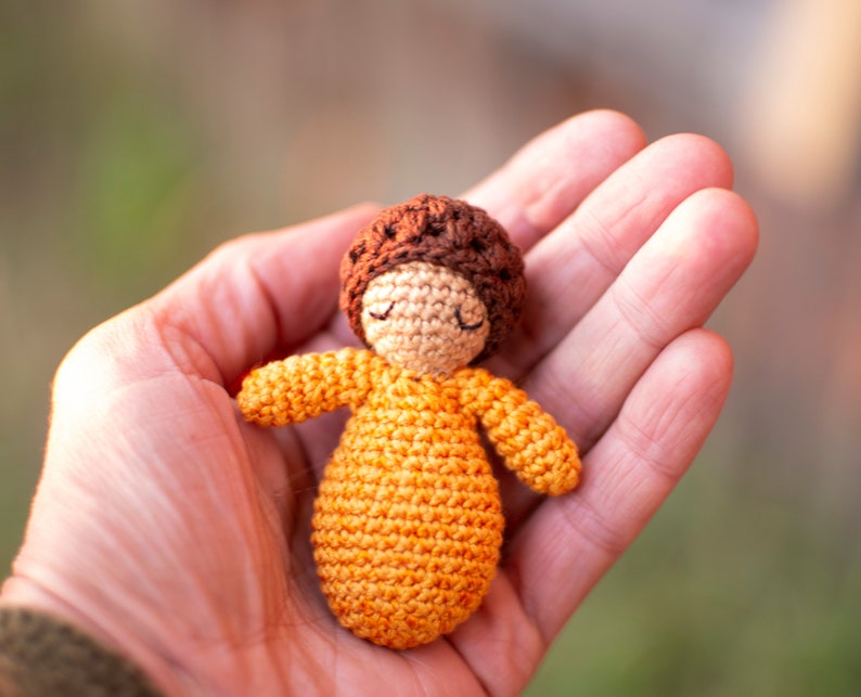 May include: A small crocheted doll with brown hair and a yellow dress. The doll is sitting in the palm of a hand.