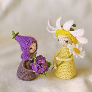 May include: Two crocheted dolls, one wearing a purple hat and holding a bouquet of purple flowers, the other wearing a white daisy flower crown and holding a white daisy.