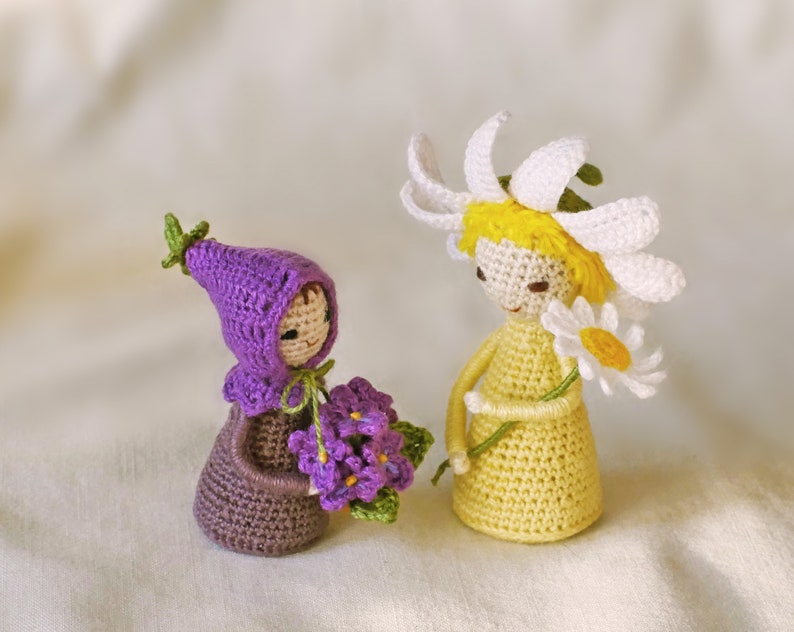 May include: Two crocheted dolls, one wearing a purple hat and holding a bouquet of purple flowers, the other wearing a white daisy flower crown and holding a white daisy.