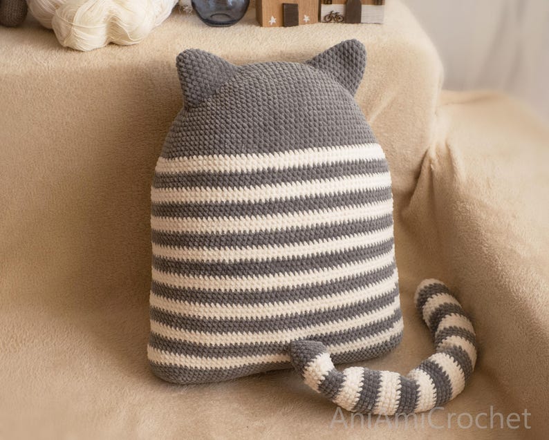May include: Gray and white striped crocheted cat pillow with a long tail. The pillow has a gray crocheted head with cat ears.