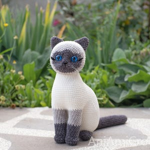 May include: A crocheted Siamese cat figurine featuring a cream-colored body, dark gray ears, face mask, paws, and tail. The cat has bright blue plastic eyes, stitched whiskers, and a seated posture. The text AniAmiCrochet is visible at the bottom.