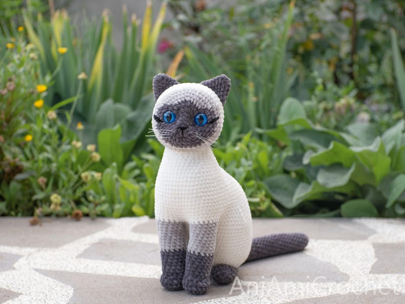 May include: A crocheted Siamese cat figurine featuring a cream-colored body, dark gray ears, face mask, paws, and tail. The cat has bright blue plastic eyes, stitched whiskers, and a seated posture. The text AniAmiCrochet is visible at the bottom.