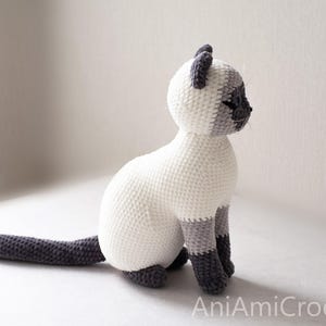 May include: A crocheted Siamese cat figurine featuring a cream-colored body with dark gray ears, face, paws, and a long tail. The cat is in a seated position with its head turned to the side. The text AniAmiCrochet is visible in the bottom corner.
