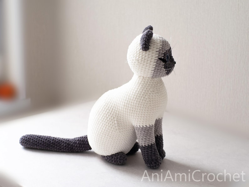 May include: A crocheted Siamese cat figurine featuring a cream-colored body with dark gray ears, face, paws, and a long tail. The cat is in a seated position with its head turned to the side. The text AniAmiCrochet is visible in the bottom corner.