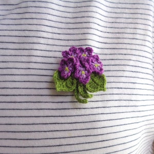May include: A crocheted flower brooch with purple flowers and green leaves.