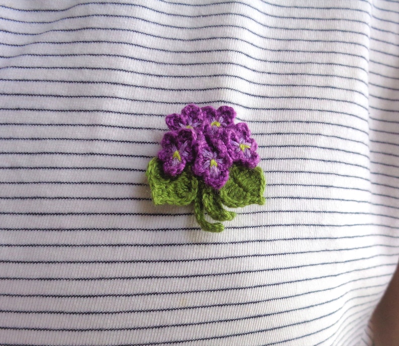 May include: A crocheted flower brooch with purple flowers and green leaves.