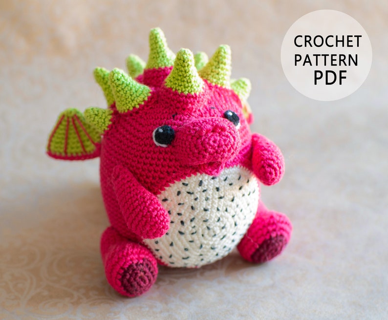 Op de afbeelding: Een gehaakt amigurumi draken speelgoed van roze en wit garen. De draak heeft groene stekels en een wit drakenfruit patroon op zijn buik. CROCHET PATTERN PDF.