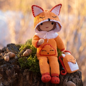 May include: A crocheted fox doll wearing an orange jumpsuit and a fox-shaped hat. The doll is sitting on a tree stump with acorns around it.