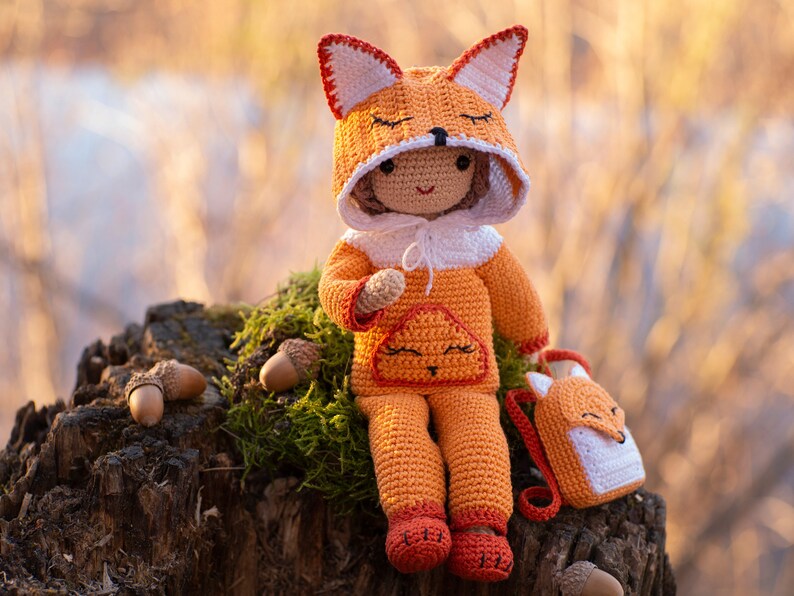 May include: A crocheted fox doll wearing an orange jumpsuit and a fox-shaped hat. The doll is sitting on a tree stump with acorns around it.